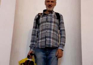 lekko uśmiechający się mężczyzna w średnim wieku ubrany w kraciastą koszulę i plecak, trzyma reklamówkę, na jasnym tle ozdobnego wgłębienia w ścianie