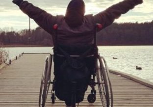 mężczyzna na wózku inwalidzkim odwrócony poecami do osoby robiącej zdjęcie i przodem do jeziora